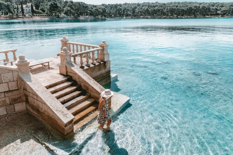 A woman walking along a stone staircase by a serene blue lake with a town in the background, Mali Losinj